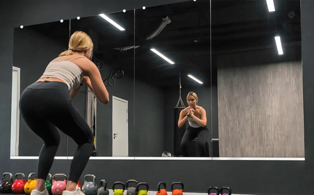 Woman doing squats in front of full-wall gym mirror with kettlebells arranged on the gym floor.