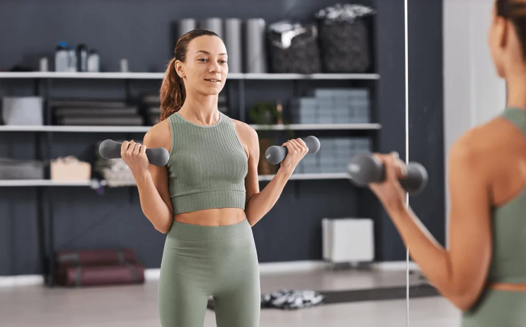 Fit woman in green activewear lifting dumbbells while checking her form in large gym mirror tiles inside a modern fitness studio.