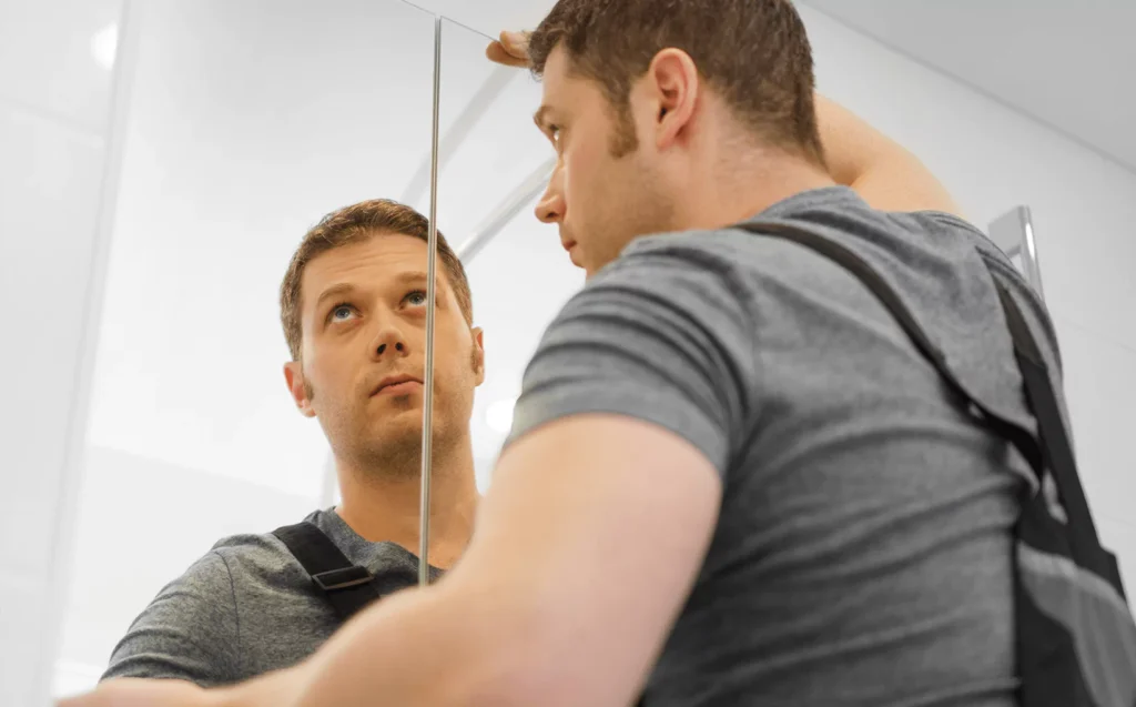 Man in gray shirt closely inspecting his reflection in a wall mirror, adjusting or evaluating positioning.