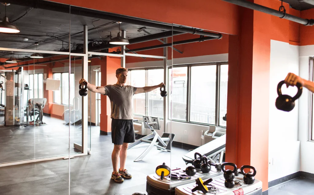 Man lifting kettlebells in front of full wall gym mirror with red and black workout room interior.