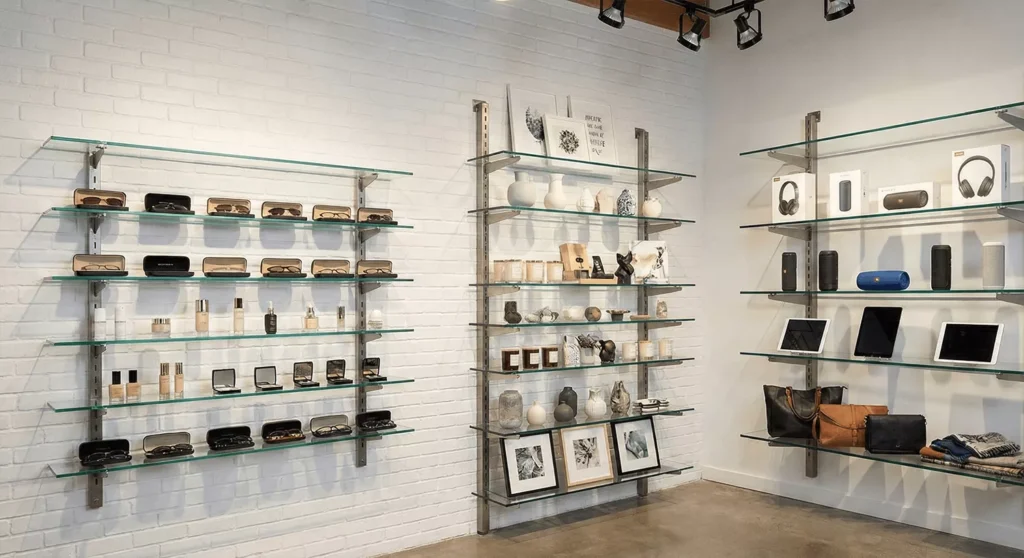 Retail glass shelves of varying depths displaying cosmetics, electronics, eyewear, and boutique items with reflections.