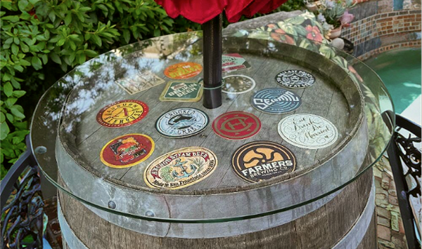 Round glass top for wine barrel table with stickers under glass, set on a wooden barrel outdoors with greenery